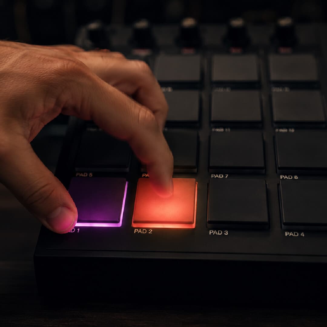 A hand pressing two adjacent pads at once on an MPC; thumb on pad 1 lit purple and index finger on pad 2 lit orange, with the rest of the grid dark.